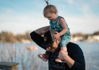 Father carrying daughter on shoulders by the lake, sharing a joyful moment together.