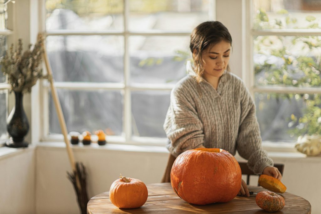 A woman in a sweater carves a pumpkin indoors, capturing autumn vibes.