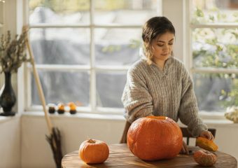 A woman in a sweater carves a pumpkin indoors, capturing autumn vibes.